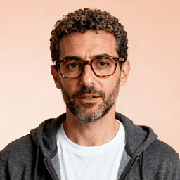 AI-generated portrait of a middle-aged man with curly hair, a beard, and glasses, wearing a hoodie over a t-shirt, posed with a relaxed expression against a neutral background in even light.