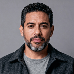 AI-generated portrait of a middle-aged man with short hair and a trimmed beard, wearing a dark jacket, posed with a serious expression against a concrete-toned background in soft moody light.