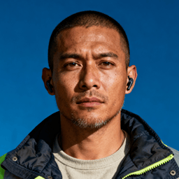 AI-generated portrait of a young man with close-cropped hair and light facial hair, wearing a work jacket, posed with a neutral expression against a dark industrial background with hard lighting.