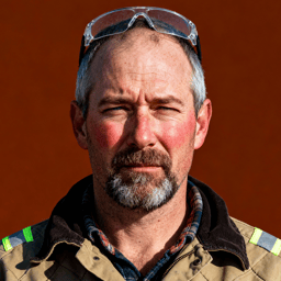 AI-generated portrait of a middle-aged man with short hair and a graying beard, wearing a rugged work jacket, posed with a tired expression against a dark industrial background with hard lighting.