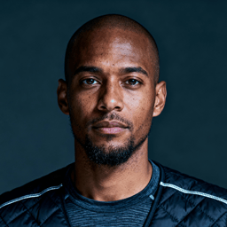 AI-generated portrait of a young man with short hair and trimmed facial hair, wearing a performance jacket, posed with a neutral expression against a dark industrial background with hard lighting.