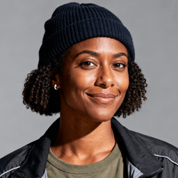 AI-generated portrait of a young woman with short curly hair under a knit beanie, wearing a utility jacket, posed with a subtle smile against a muted background with hard lighting.