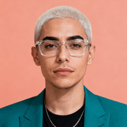 AI-generated portrait of a young person with closely cropped hair and glasses, wearing a blazer and necklace, posed with a neutral expression against a coral background in high-key lighting.