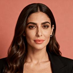 AI-generated portrait of a young woman with long wavy hair and defined brows, wearing a tailored blazer, posed with a confident expression against a coral background in balanced lighting.