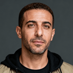 AI-generated portrait of a middle-aged man with short hair and light stubble, wearing a hoodie under a jacket, posed with a neutral expression against a dark gray background in soft studio lighting.