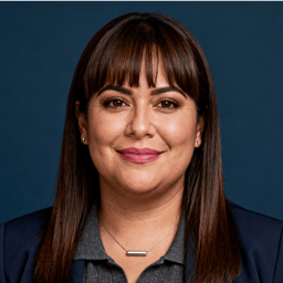 AI-generated portrait of a middle-aged woman with straight hair and bangs, wearing a collared top and blazer, posed with a subtle smile against a dark blue background in soft studio lighting.