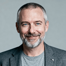 AI-generated portrait of a middle-aged man with short gray hair and a trimmed beard, wearing a blazer over a t-shirt, posed with a relaxed smile against a light gray background in soft studio lighting.