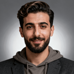 AI-generated portrait of a young man with short styled hair and a trimmed beard, wearing a hoodie under a blazer, posed with a gentle smile against a soft gray background in clean studio lighting.