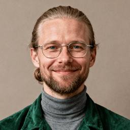 AI-generated portrait of a middle-aged man with long hair pulled back and glasses, wearing a turtleneck and jacket, posed with a gentle smile against a neutral beige background in soft studio light.