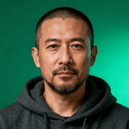 AI-generated portrait of a middle-aged man with closely cropped hair and a short beard, wearing a hoodie, posed with a neutral expression against a green background in cool studio lighting.
