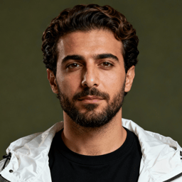 AI-generated portrait of a young man with wavy hair and a full beard, wearing a jacket over a t-shirt, posed with a calm expression against an olive green background in balanced light.
