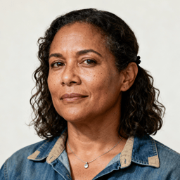 AI-generated portrait of a middle-aged woman with shoulder-length curly hair and light freckles, wearing a denim shirt and necklace, posed with a gentle expression against an off-white background in natural light.