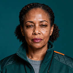 AI-generated portrait of a middle-aged woman with tightly curled hair and faint freckles, wearing a quilted utility jacket, posed with a serious expression against a teal-toned background in high-contrast lighting.