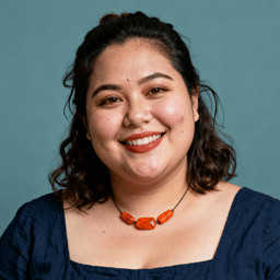 AI-generated portrait of a middle-aged woman with wavy hair pulled back, wearing a simple linen top and necklace, posed with a friendly smile against a muted teal background in diffused light.