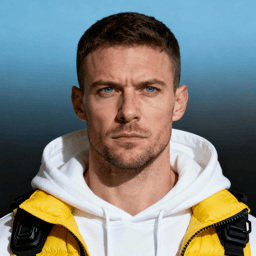 AI-generated portrait of a young man with short hair and light stubble, wearing a yellow technical jacket over a white hoodie, posed with a serious expression against a blue gradient background in directional lighting.
