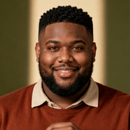 AI-generated portrait of a young man with short textured hair and a trimmed beard, wearing a collared shirt and knit sweater, posed with a soft smile against an olive background in diffused light.