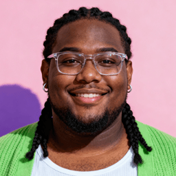 AI-generated portrait of a young man with braided hair and glasses, wearing a green cardigan and white shirt, posed with a gentle smile against a pink background in soft cool light.