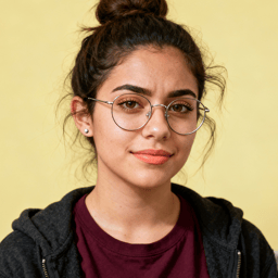 AI-generated portrait of a young woman with dark hair in a messy bun and round glasses, wearing a hoodie over a T-shirt, posed with a neutral expression against a warm yellow indoor background in soft light.