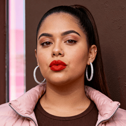 AI-generated portrait of a young woman with long pulled-back hair and defined makeup, wearing hoop earrings and a padded jacket, posed with a confident expression against a dark studio background in single-source light.