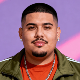 AI-generated portrait of a young man with a close-cropped haircut and trimmed goatee, wearing an olive jacket and orange shirt, posed with a neutral expression against a pastel background in bright studio light.