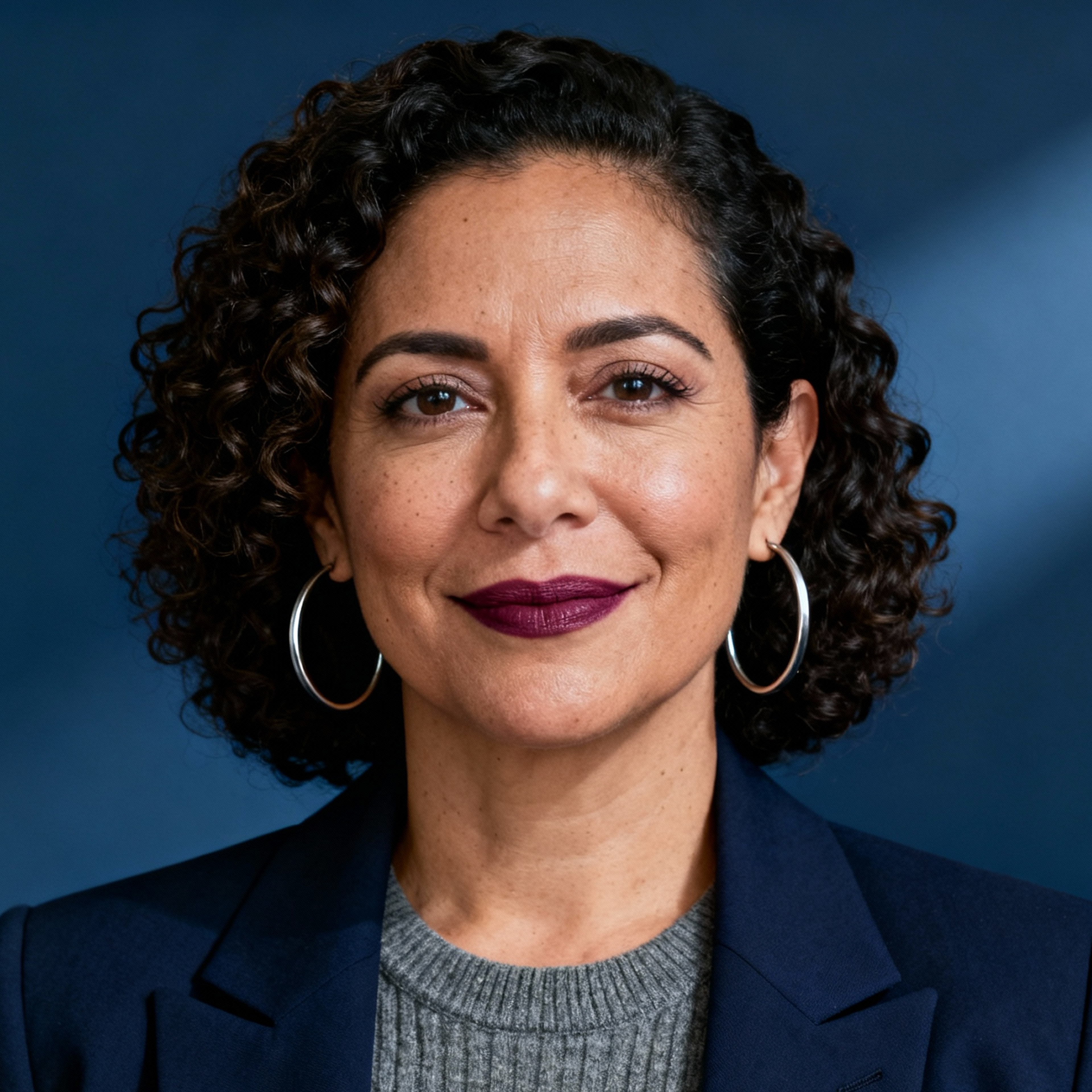 AI-generated portrait of a middle-aged woman with short curly hair and defined features, wearing a blazer and knit top, posed with a confident expression against a muted blue background in soft studio lighting.